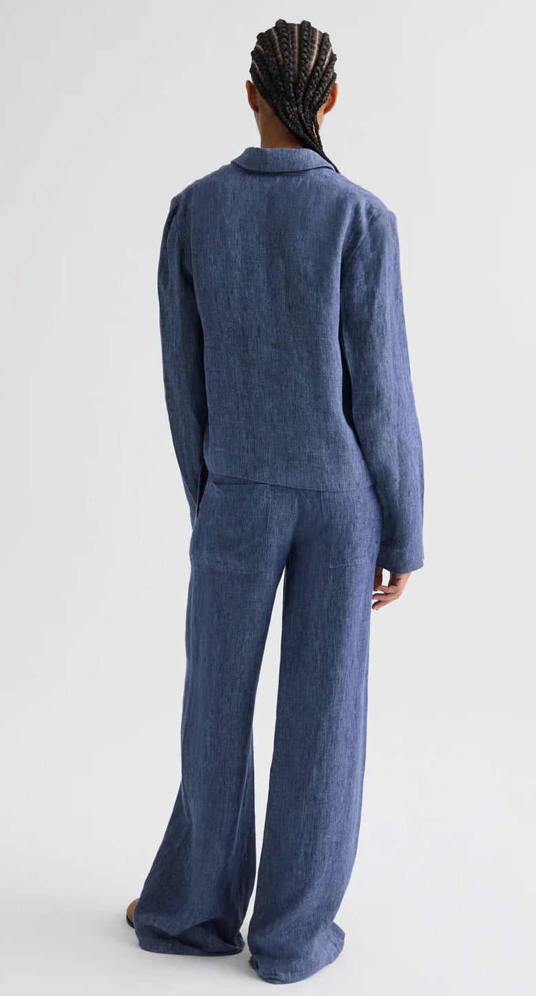 Person with back to camera wears the TWP Lucy Hemp Denim Top in blue—a loose, boxy silhouette with long sleeves—paired with wide-leg pants against a plain light background.