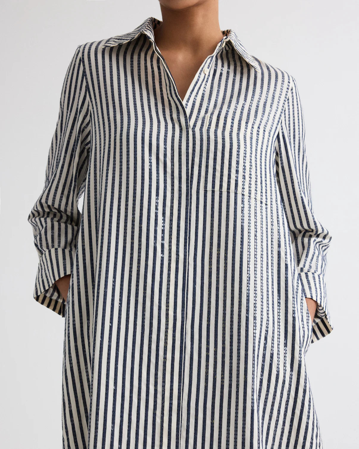 Person wearing TWP’s Jenny's Clear Sequin Stripe Gown, featuring vertical navy and white stripes and crafted from Italian cotton shirting, photographed from the shoulders down with hands in pockets.