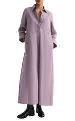 A person wears TWP's Jenny's Framed Micro Striped Gown—a long, loose-fitting, light purple striped maxi shirt dress with a collar and chest pocket—crafted from Italian cotton shirting, paired with black ankle boots.