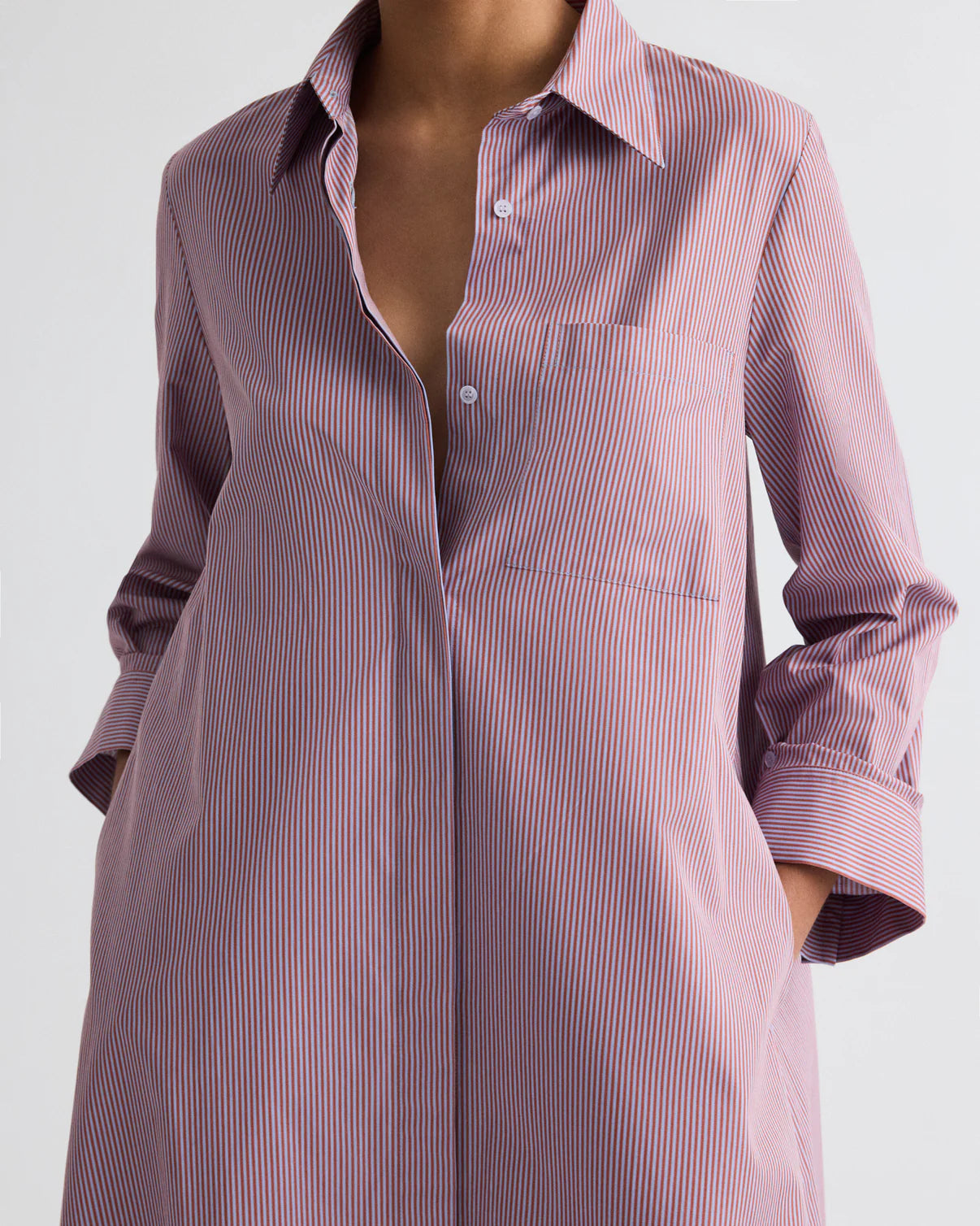 A person wears the TWP Jenny’s Framed Micro Striped Gown, an Italian cotton shirting maxi dress with an A-line silhouette and thin red and blue vertical stripes. Hands rest in pockets; face not visible.