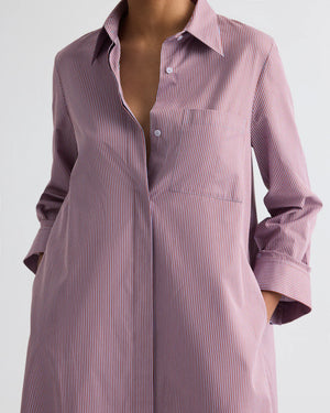 A person wears the TWP Jenny’s Framed Micro Striped Gown, an Italian cotton shirting maxi dress with an A-line silhouette and thin red and blue vertical stripes. Hands rest in pockets; face not visible.