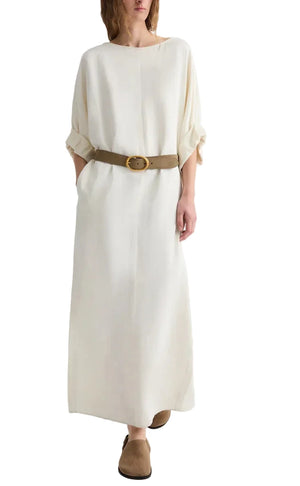 A person is wearing the TWP Dress for B, a cream-colored European flax linen maxi dress with a boat neck and rolled sleeves, styled with a brown belt and brown shoes, standing against a white background.
