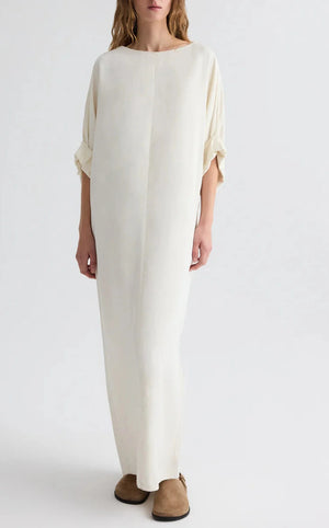 A person in the TWP Dress for B—a loose, off-white boat neck maxi dress crafted from European flax linen with rolled sleeves—pairs it with brown slippers and stands against a plain light background.