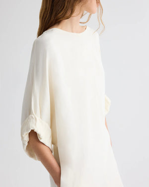 A woman stands against a plain background wearing the TWP Dress for B, a loose-fitting European flax linen blouse with elbow-length sleeves and her hands in her pockets.