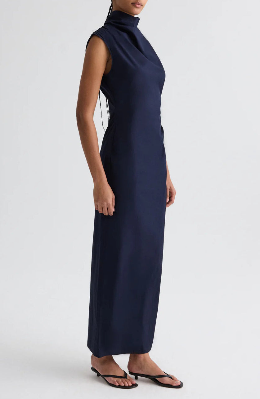 A model wears the TWP Gathered Thoughts Dress, a sleeveless navy blue maxi with a high neck, paired with black sandals, set against a plain white background.