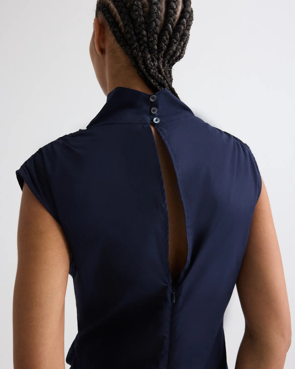 A person with braided hair wears the Gathered Thoughts Dress by TWP—a navy cap-sleeve dress with a back slit and two collar buttons, evoking the elegance of an open-back style.