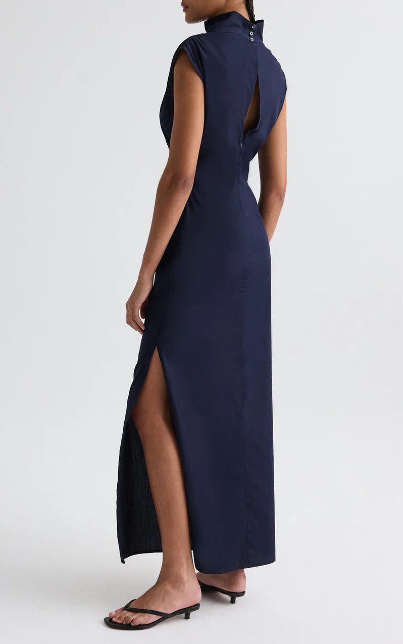 A person models the TWP Gathered Thoughts Dress in navy blue, featuring an open back, side slit, and keyhole detail, paired with black sandals against a plain light background.