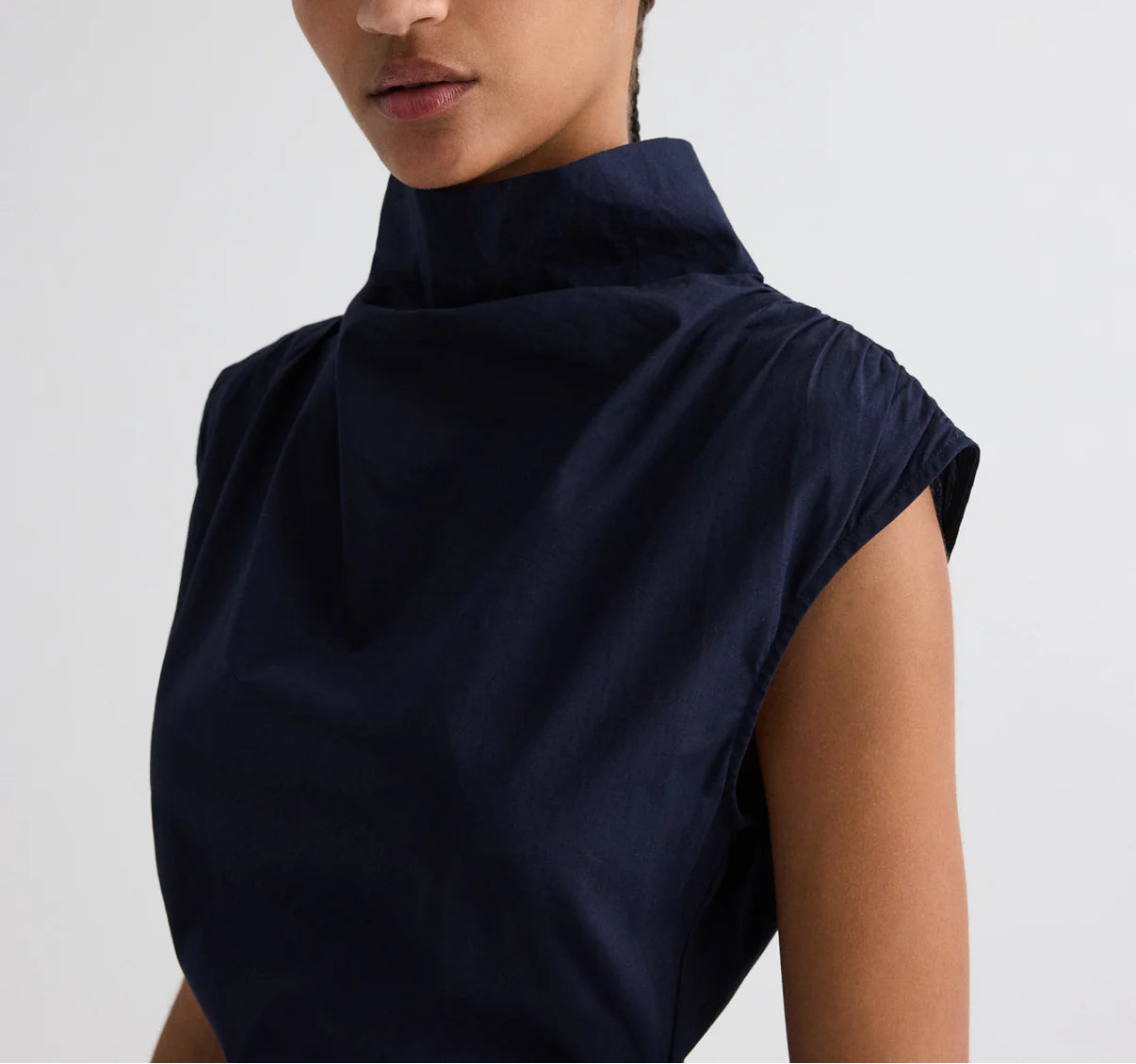A person wears the Gathered Thoughts Dress by TWP, a sleeveless, high-neck navy blue stretch cotton piece, shown from the shoulders up against a plain background.