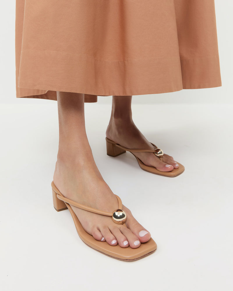 A person wearing a tan midi skirt and Loeffler Randall’s Odessa Heeled Mule in beige nappa leather with a gold ball detail, standing on a white background.
