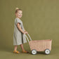 Toddler girl in a gray dress and yellow sandals, smiling and pulling an Olli Ella Rattan Strolley against a green backdrop.