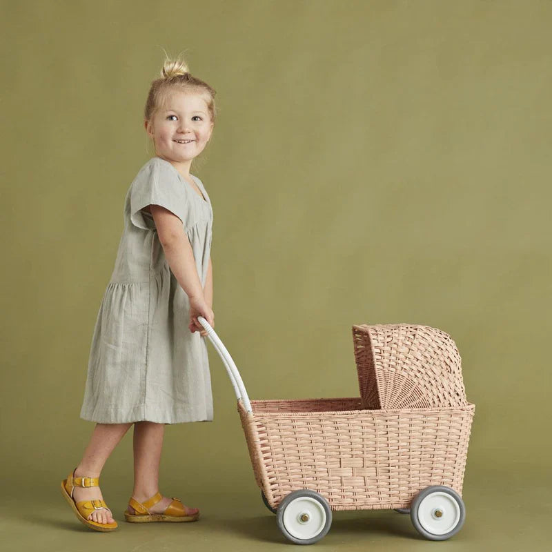 Young girl in a gray dress and yellow sandals, smiling and pulling an Olli Ella Rattan Strolley, standing against a green backdrop.