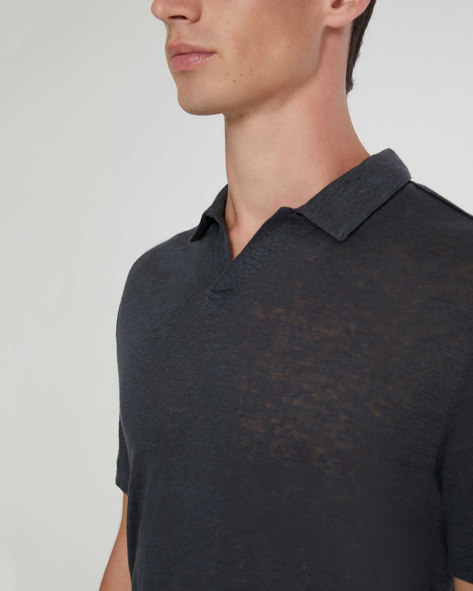A man models the Onia Shaun Linen Polo, a dark linen jersey with a relaxed fit and split Johnny collar, shown from the shoulders up against a plain background.