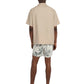 Facing away, a person wears Onia's Charles 5" Swim Trunk in white with green leaf designs and a beige short-sleeve shirt. The quick-dry swim trunks stand out against the plain background as they remain barefoot.
