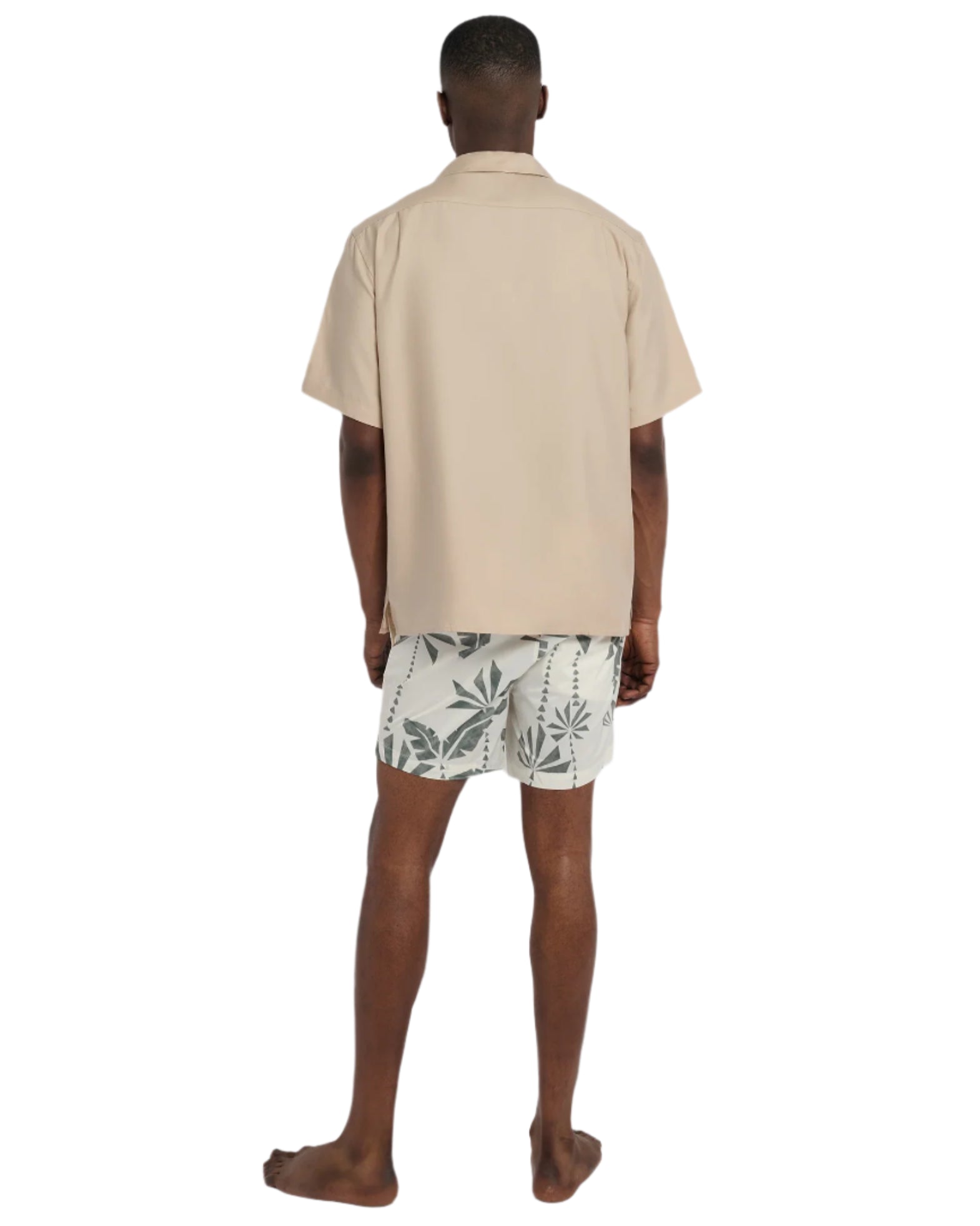 Facing away, a person wears Onia's Charles 5" Swim Trunk in white with green leaf designs and a beige short-sleeve shirt. The quick-dry swim trunks stand out against the plain background as they remain barefoot.