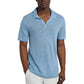 A man wears the Onia Shaun Linen Polo in light blue, featuring a relaxed fit and Johnny collar, paired with white pants as he stands against a plain white background with one hand in his pocket.