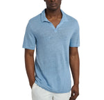 A man wears the Onia Shaun Linen Polo in light blue, featuring a relaxed fit and Johnny collar, paired with white pants as he stands against a plain white background with one hand in his pocket.