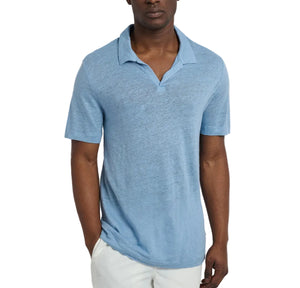 A man wears the Onia Shaun Linen Polo in light blue, featuring a relaxed fit and Johnny collar, paired with white pants as he stands against a plain white background with one hand in his pocket.