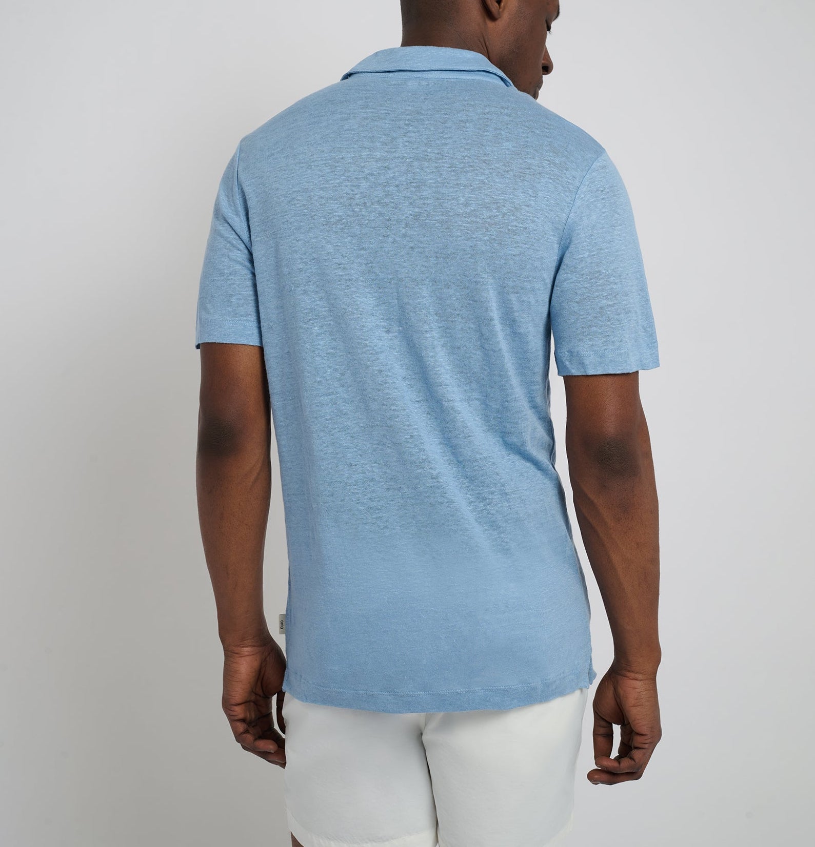 A man stands with his back to the camera, wearing the Onia Shaun Linen Polo in light blue and white shorts against a plain background.