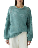 A person wearing the Guest in Residence Open Stripe Crew, a lightweight striped sweater in light teal, paired with white pants and a chunky gold necklace, stands against a white background.