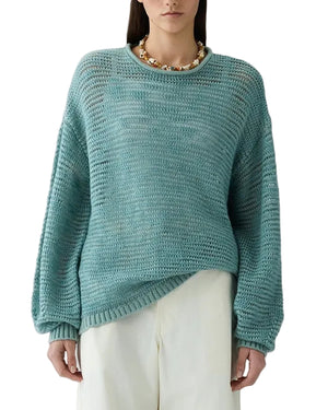 A person wearing the Guest in Residence Open Stripe Crew, a lightweight striped sweater in light teal, paired with white pants and a chunky gold necklace, stands against a white background.