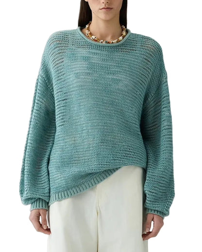 A person wearing the Guest in Residence Open Stripe Crew, a lightweight striped sweater in light teal, paired with white pants and a chunky gold necklace, stands against a white background.