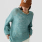 A person wearing the Open Stripe Crew sweater by Guest in Residence, styled with beige pants, a brown New York Yankees cap, and a beaded necklace stands against a plain background.