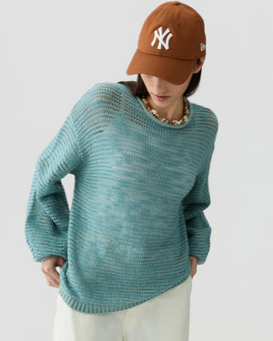 A person wearing the Open Stripe Crew sweater by Guest in Residence, styled with beige pants, a brown New York Yankees cap, and a beaded necklace stands against a plain background.