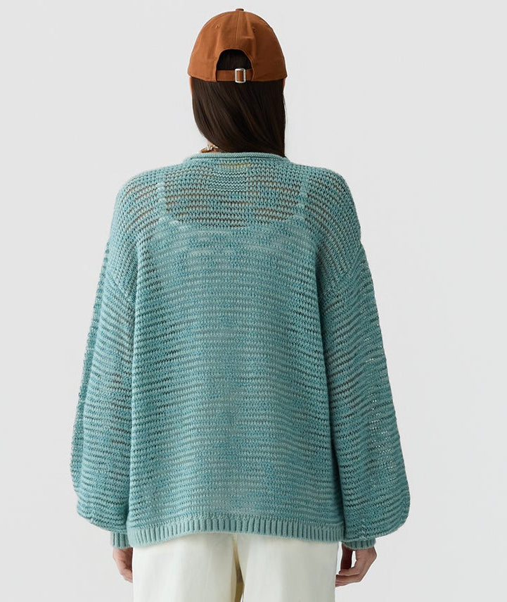 A person is shown from the back against a plain white background, wearing the Guest in Residence Open Stripe Crew—a loose, light blue merino wool sweater—paired with beige pants and a brown cap.
