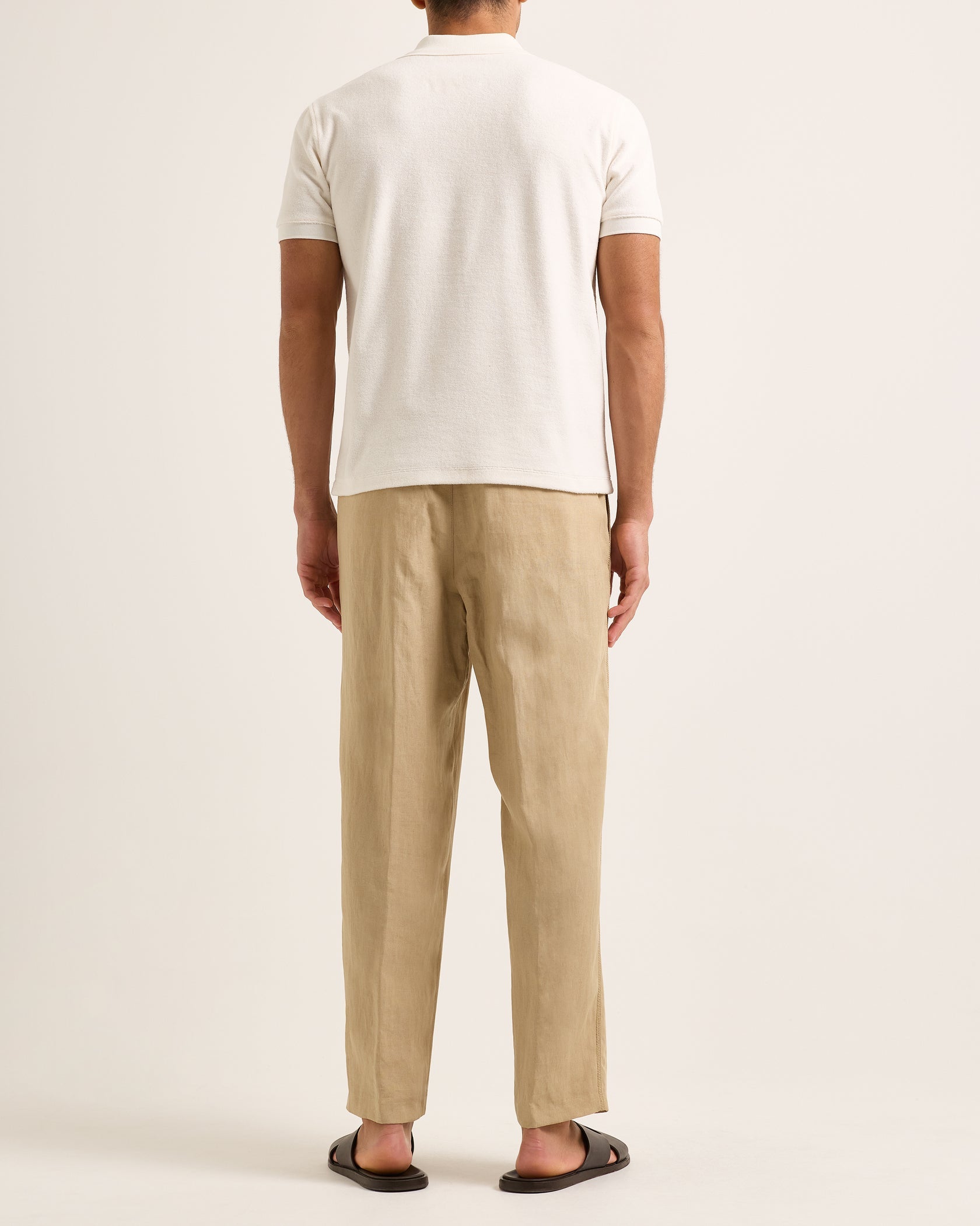 A man stands with his back to the camera wearing the Orlebar Brown Bruno Towelling Polo in white, paired with beige pants and black sandals against a plain background.