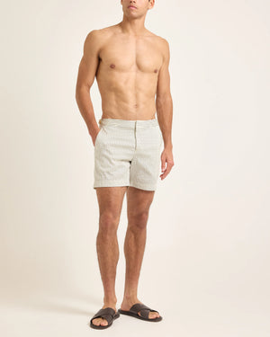 A man stands against a plain background wearing Orlebar Brown Bulldog Classic Stripe Shorts—light, quick-drying tailored swim trunks—and dark sandals, with hands in pockets and no shirt.