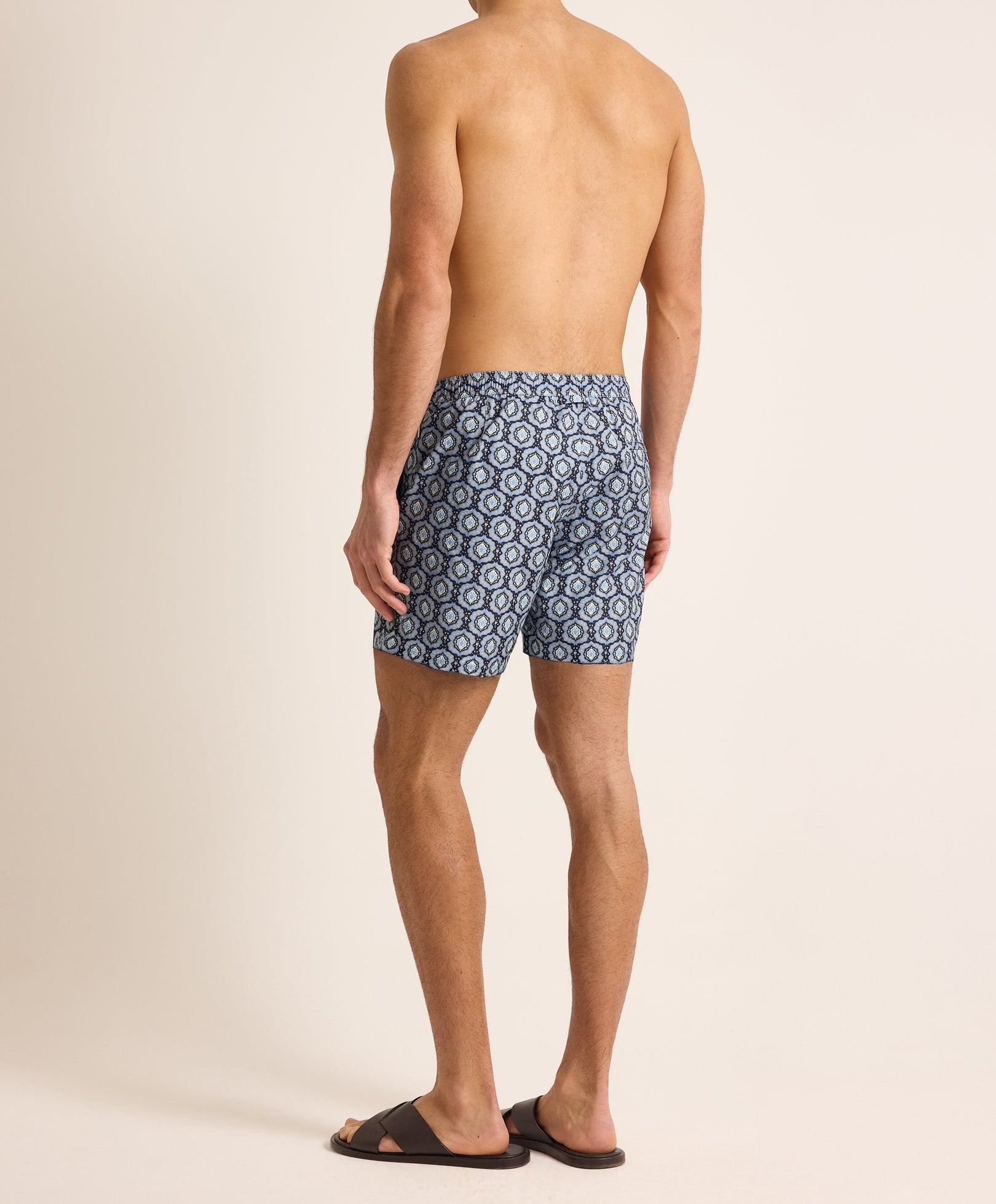 A man faces away, wearing Orlebar Brown Bulldog Drawcord Passetto Swim Trunk in a patterned blue with black slides, set against a plain background.