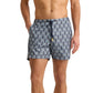 A man wears Orlebar Brown Bulldog Drawcord Passetto Swim Trunk, featuring a blue geometric pattern and made from recycled polyester, standing with one hand in his pocket.