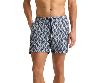 A man wears Orlebar Brown Bulldog Drawcord Passetto Swim Trunk, featuring a blue geometric pattern and made from recycled polyester, standing with one hand in his pocket.