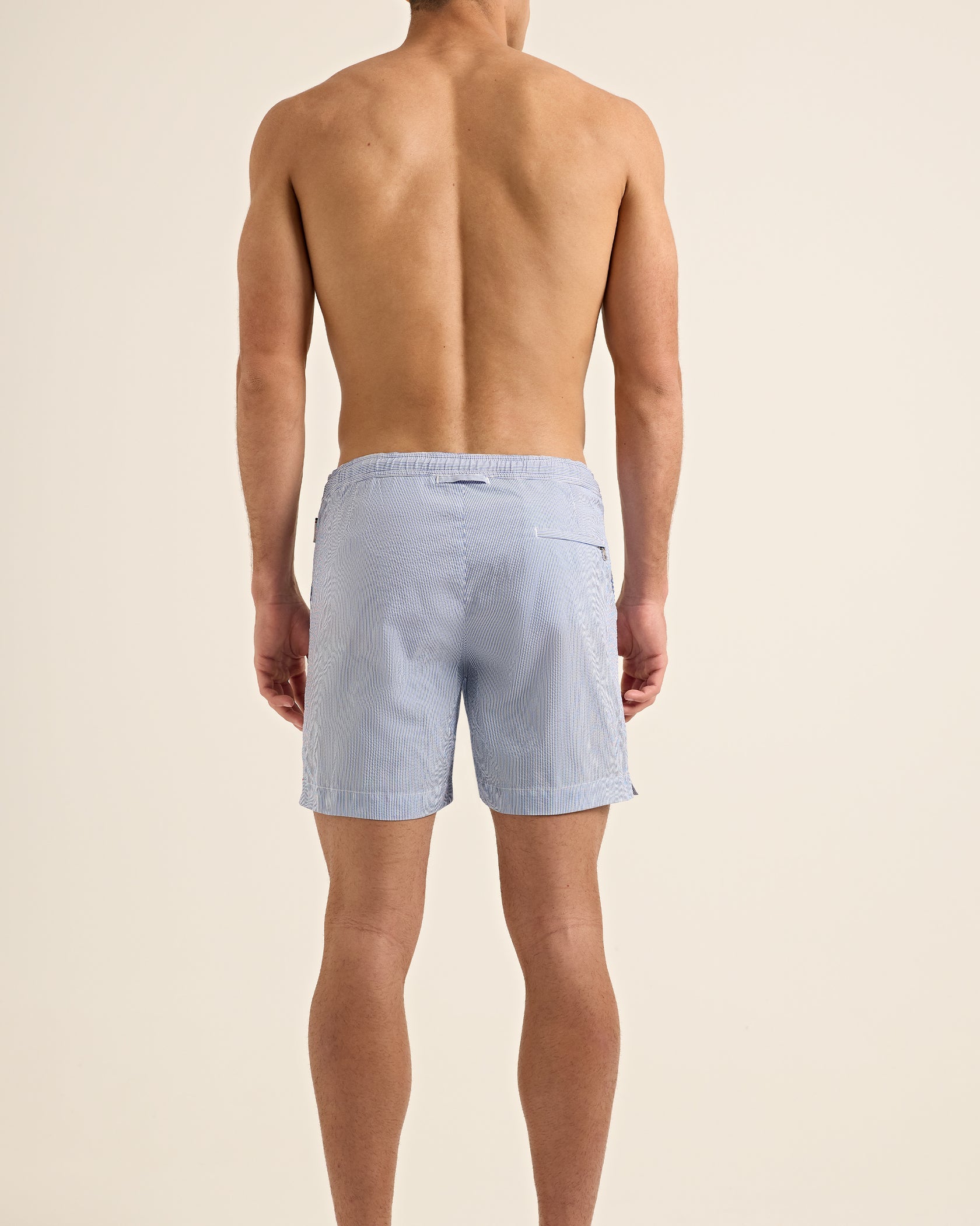 A man stands barefoot with his back to the camera, wearing Orlebar Brown Bulldog Drawcord Seersucker Shorts in light blue against a plain background.