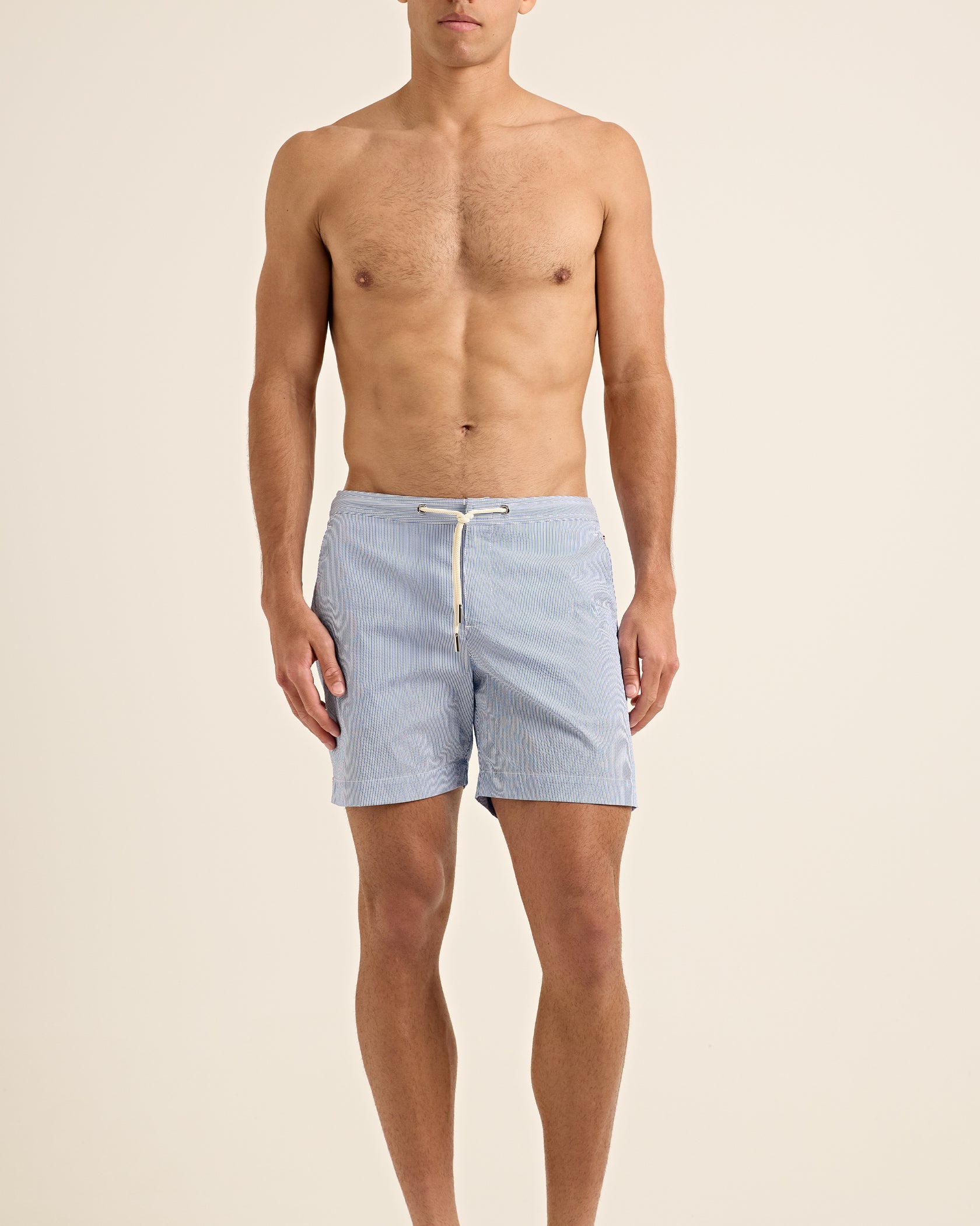 A shirtless man stands facing forward against a plain beige background, wearing Orlebar Brown's Bulldog Drawcord Seersucker Short—lightweight swim trunks with a rope-style drawcord and subtle stripes.