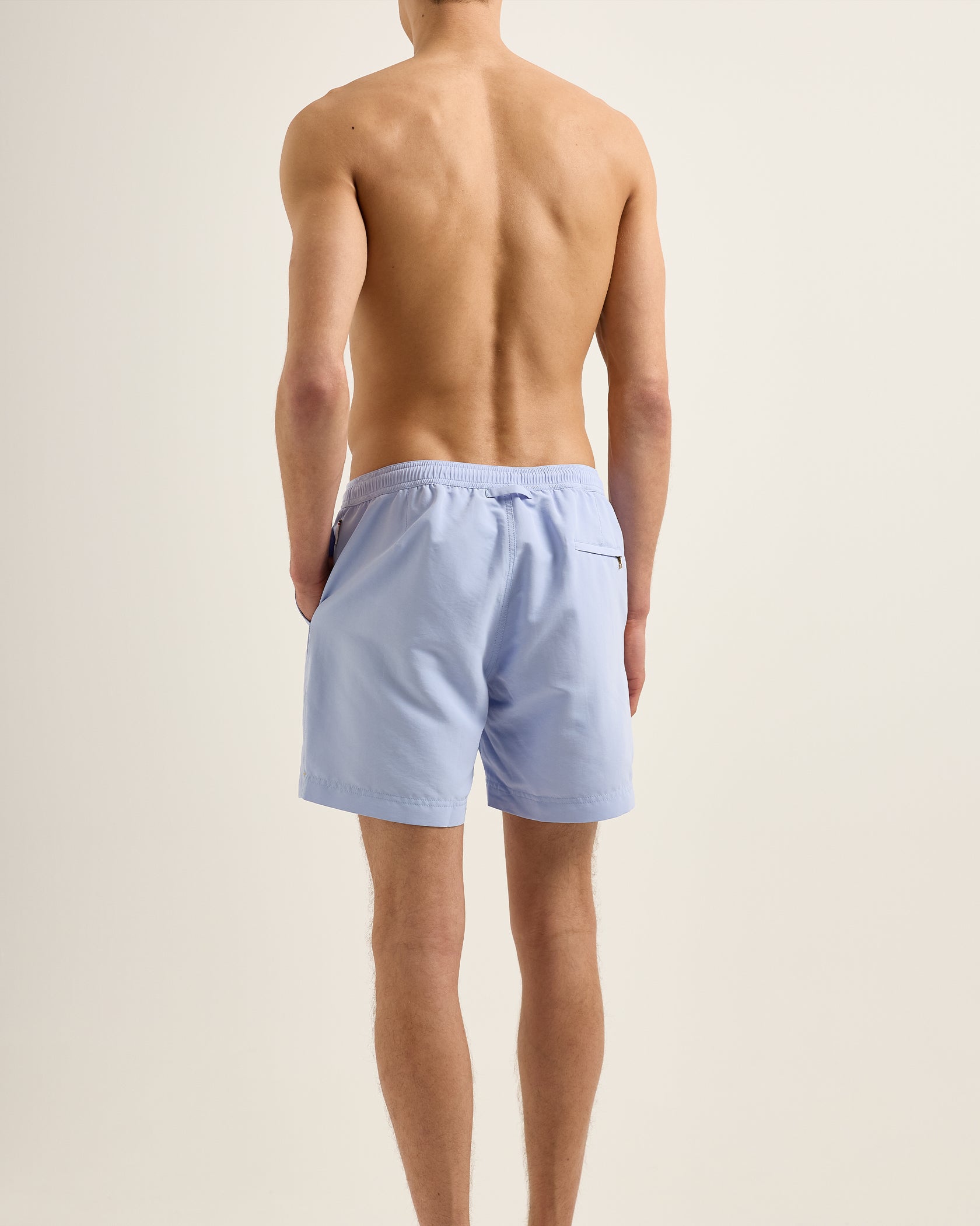 A man faces away from the camera against a plain background, wearing Orlebar Brown Bulldog Drawcord Swim Shorts in light blue with a rope-style drawcord and no shirt.