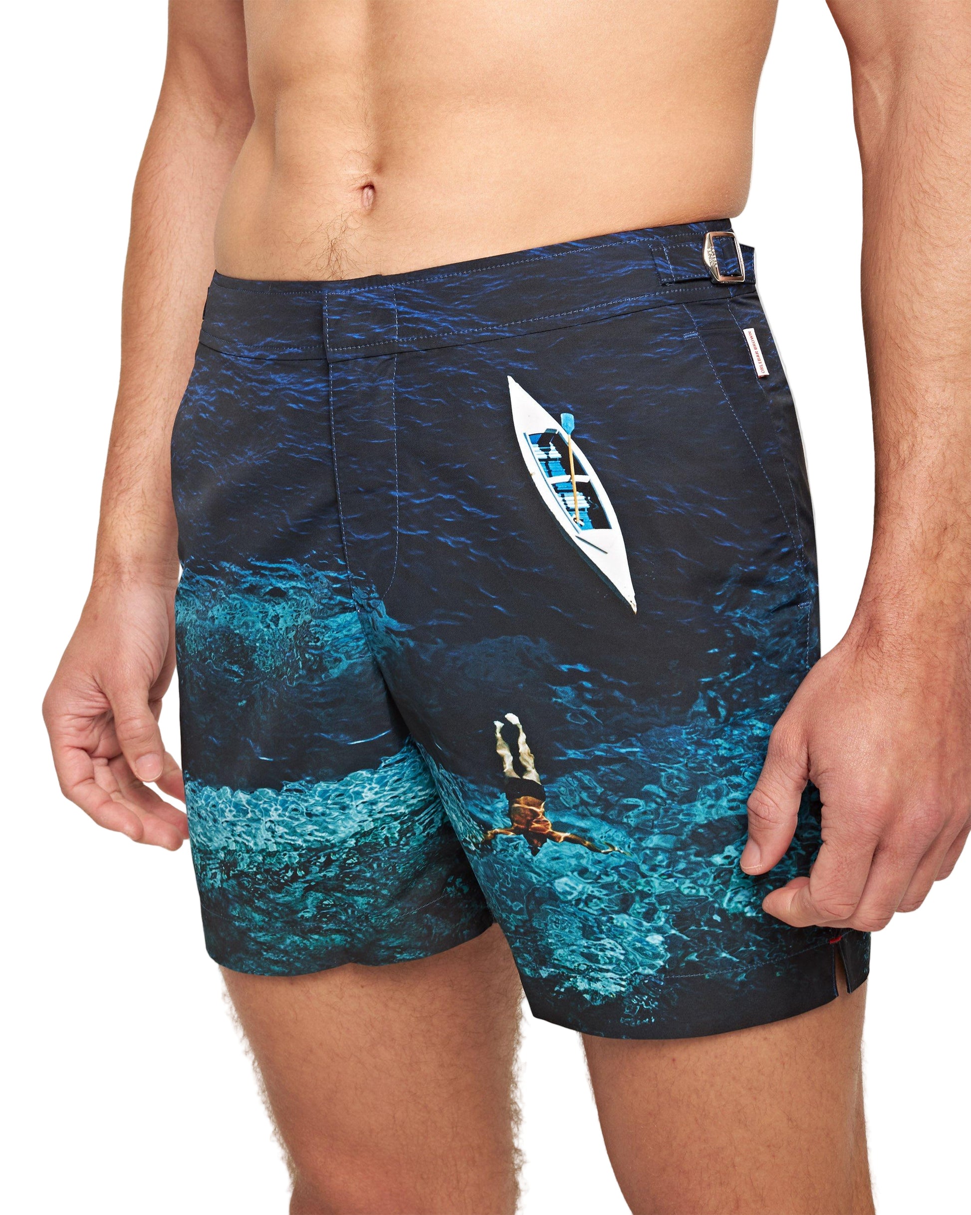 A man wearing Orlebar Brown Bulldog Photographic Swim Trunks featuring an aerial print of a swimmer diving underwater and a white kayak on blue water.
