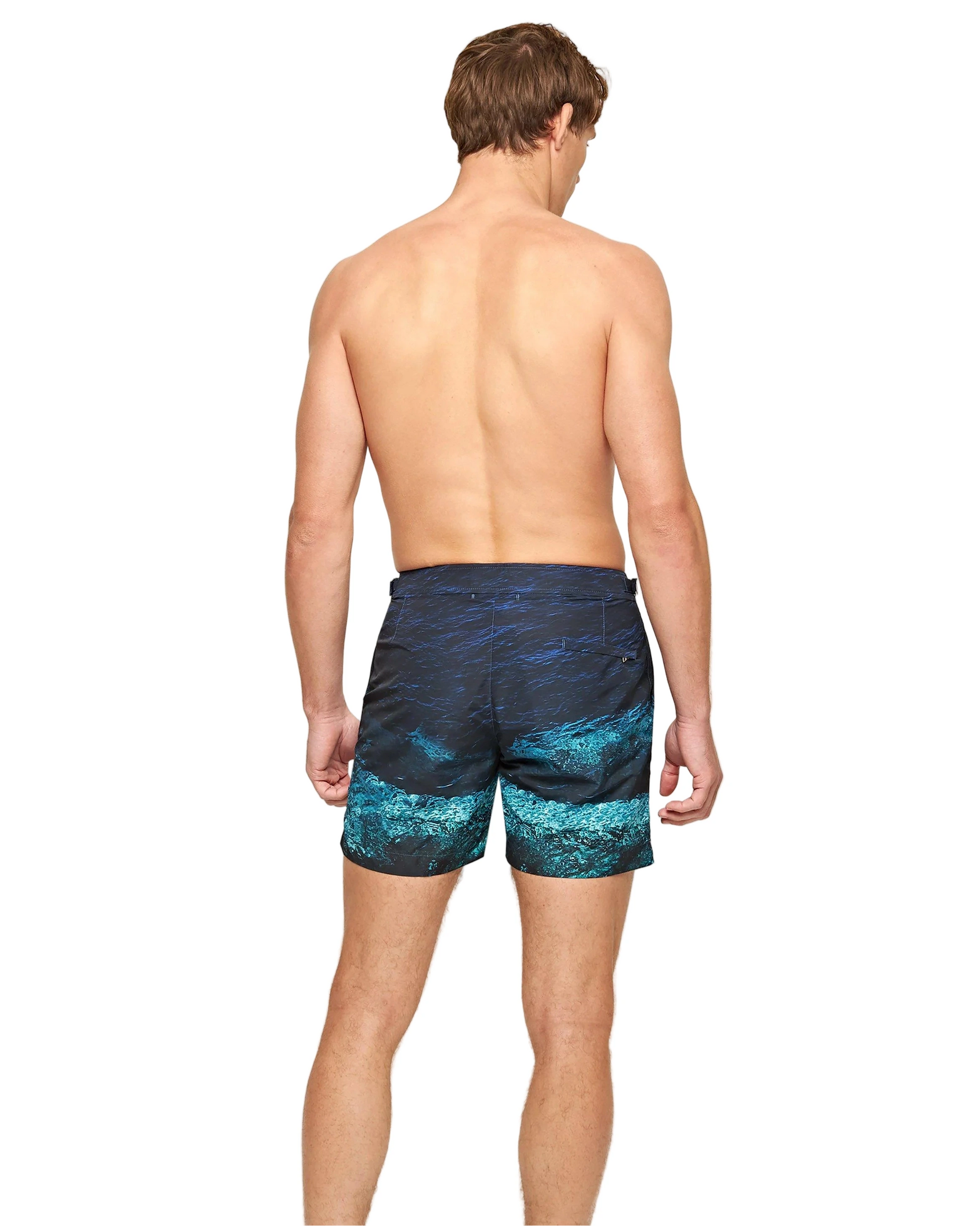 A man faces away from the camera, shirtless and barefoot, wearing Orlebar Brown Bulldog Photographic Swim Trunks featuring a blue and teal water design.