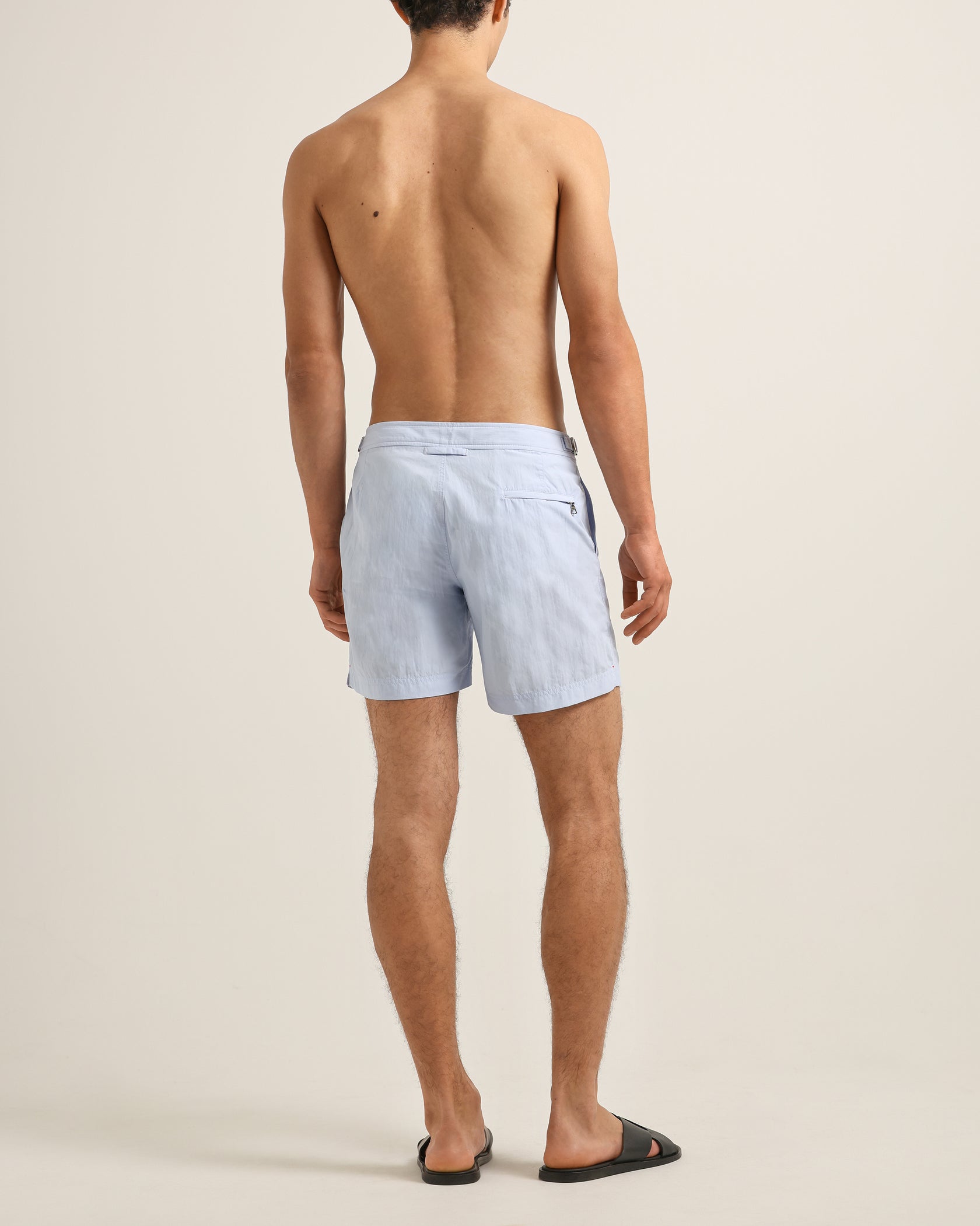 A man faces away from the camera, wearing Orlebar Brown Bulldog Swim Shorts in Surfside Blue and black slide sandals, set against a neutral background.