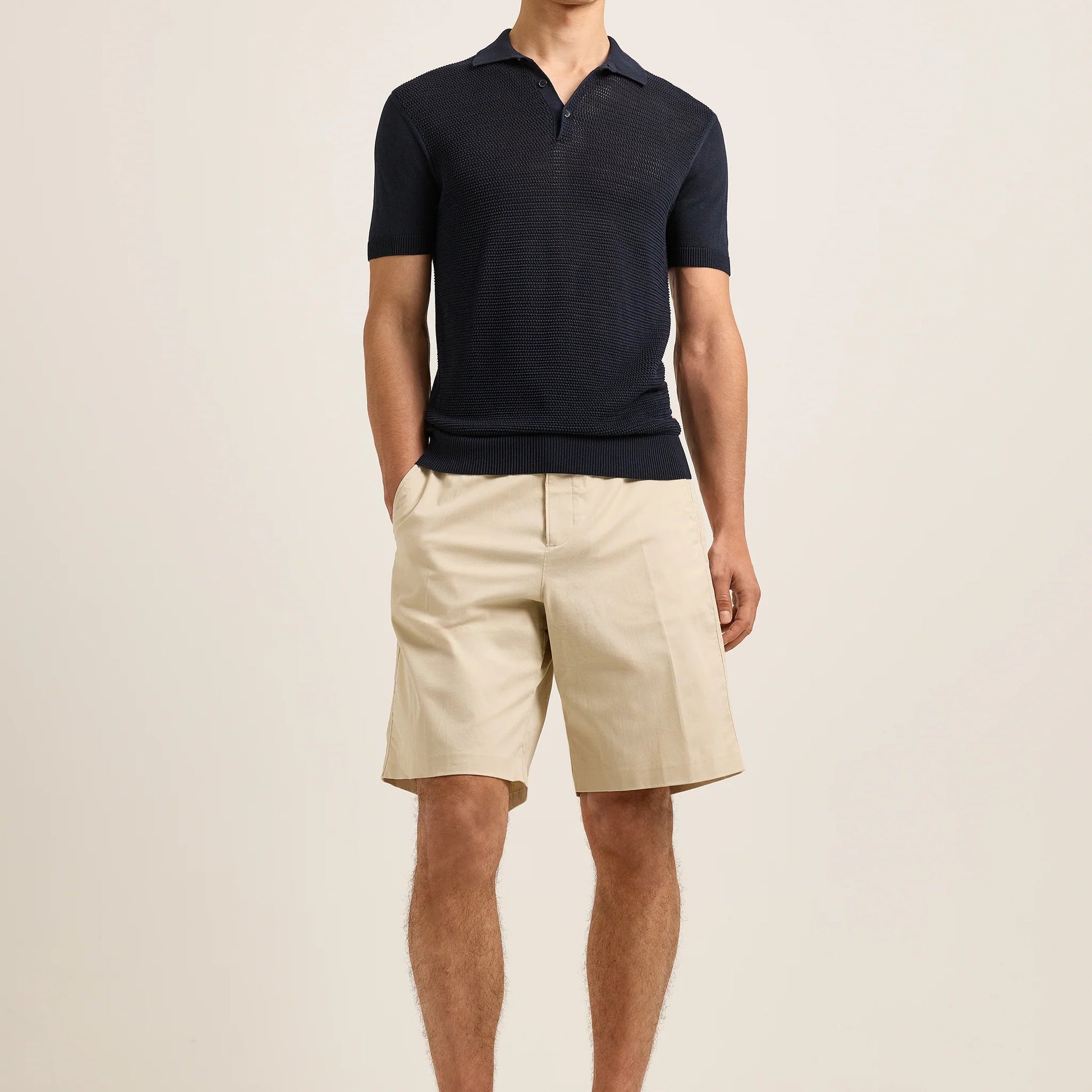 Wearing the Orlebar Brown Keeling 3 Button Polo in understated navy, paired with refined beige knee-length shorts, a person stands against a plain light background, evoking the deep elegance of Night Iris.