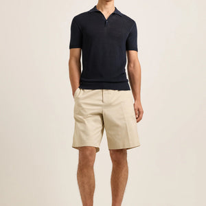 Wearing the Orlebar Brown Keeling 3 Button Polo in understated navy, paired with refined beige knee-length shorts, a person stands against a plain light background, evoking the deep elegance of Night Iris.