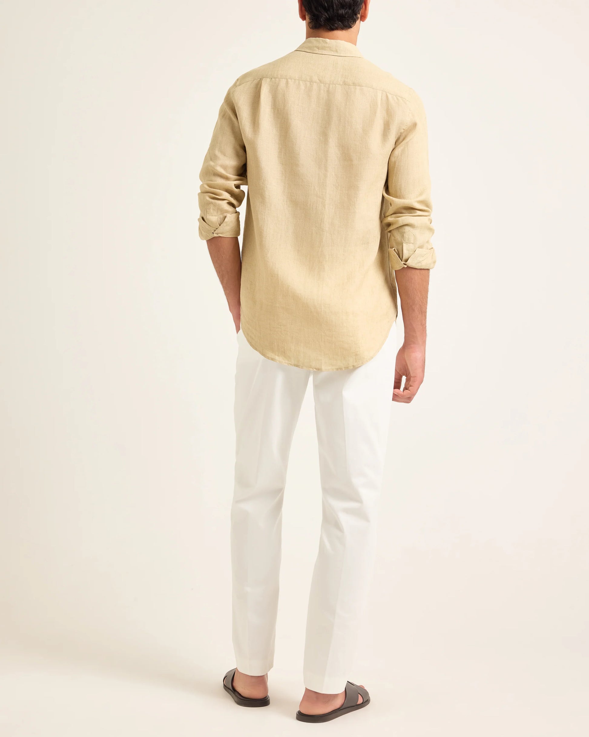 A person stands facing away, wearing the Orlebar Brown Colburn Lino Shirt in beige, paired with white trousers and dark sandals, a linen handwoven scarf draped casually over their shoulders against a plain background.