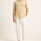 A man wearing the Orlebar Brown Colburn Lino Shirt, a beige textured linen long-sleeve button-up, with white pants and dark slide sandals stands against a plain light background.
