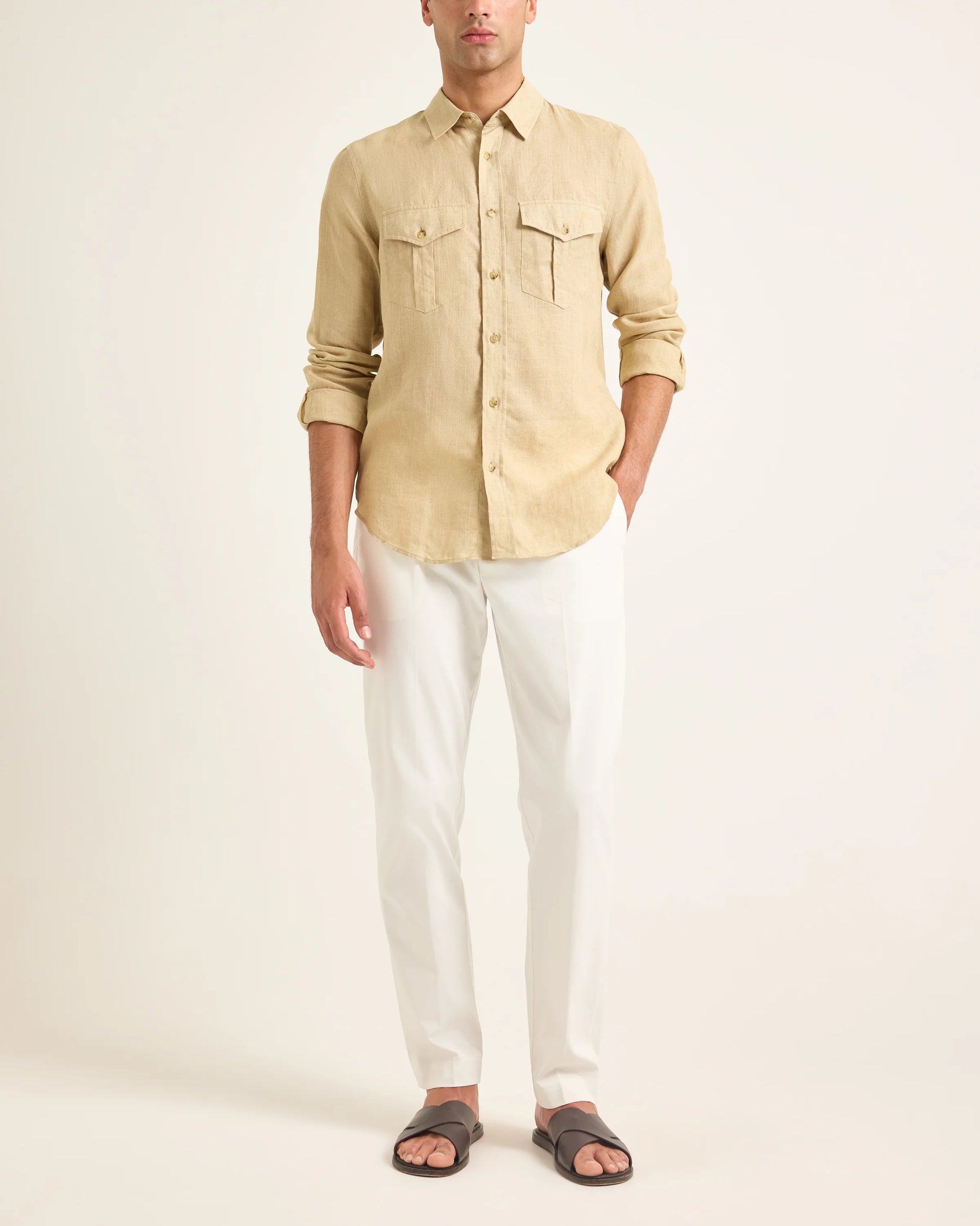 A man wearing the Orlebar Brown Colburn Lino Shirt, a beige textured linen long-sleeve button-up, with white pants and dark slide sandals stands against a plain light background.