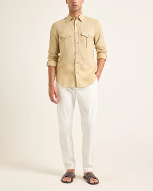 A man wearing the Orlebar Brown Colburn Lino Shirt, a beige textured linen long-sleeve button-up, with white pants and dark slide sandals stands against a plain light background.