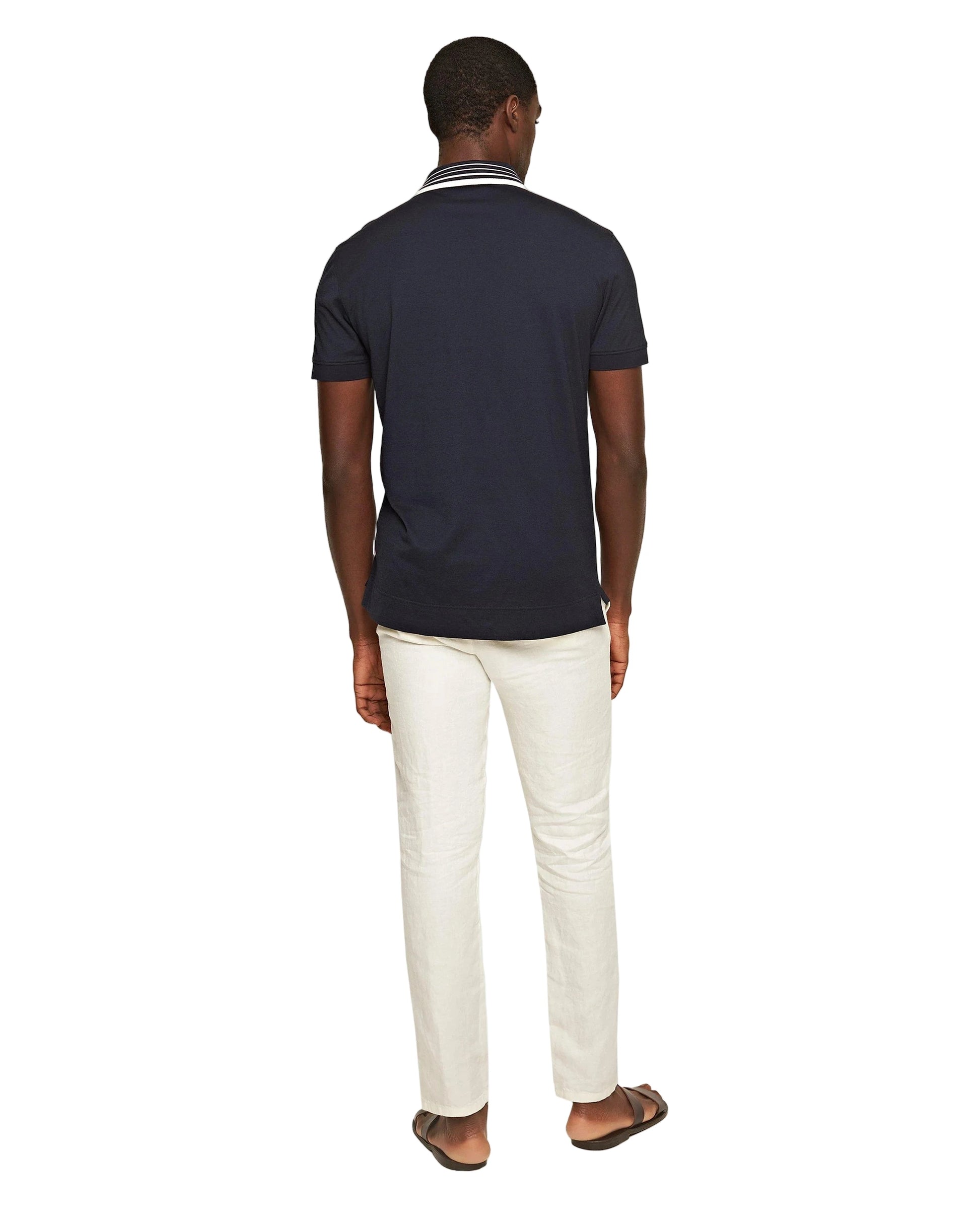 A man faces away, wearing a navy short-sleeve polo, Orlebar Brown Cornell Linen Trouser with drawcord waistband, and brown sandals against a white background.