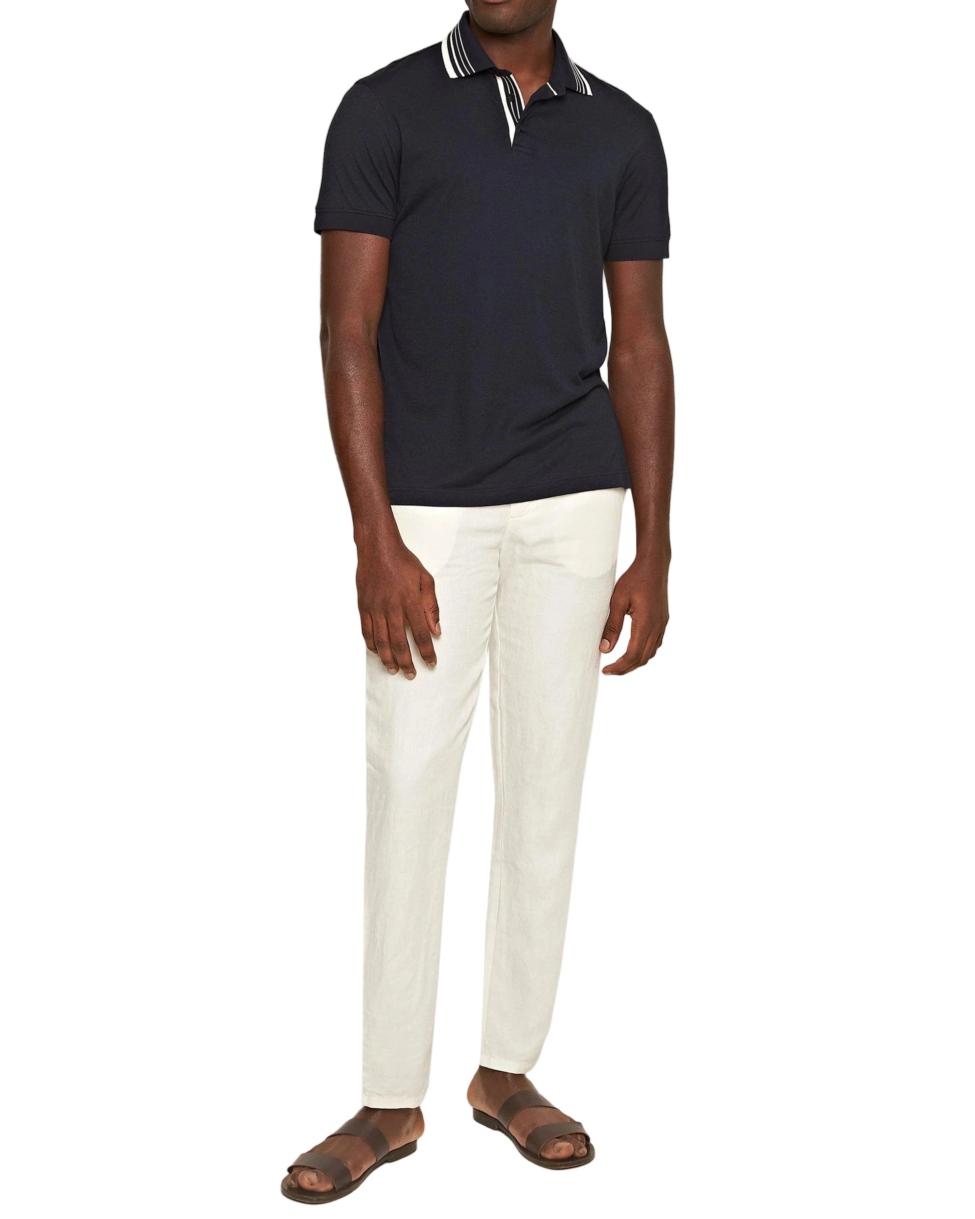 A man in a black short-sleeve polo, Orlebar Brown Cornell Linen Trousers with a drawcord waistband, and brown sandals stands against a plain white background.