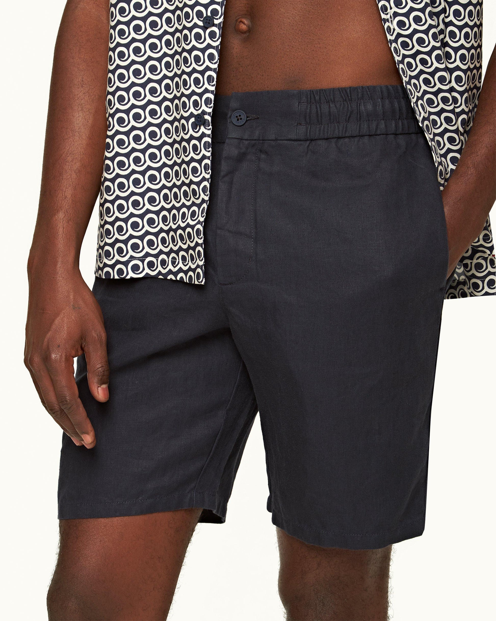 A person wears Orlebar Brown’s Cornell Linen Short—Italian-woven with an elasticated waistband—paired with a patterned shirt. The image is cropped to focus on the torso and upper legs, one hand relaxed at their side.