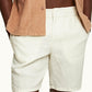 A person wears Orlebar Brown Cornell Linen Shorts in white with an elastic waistband, paired with a beige shirt, hands in pockets, shown from waist to mid-thigh.