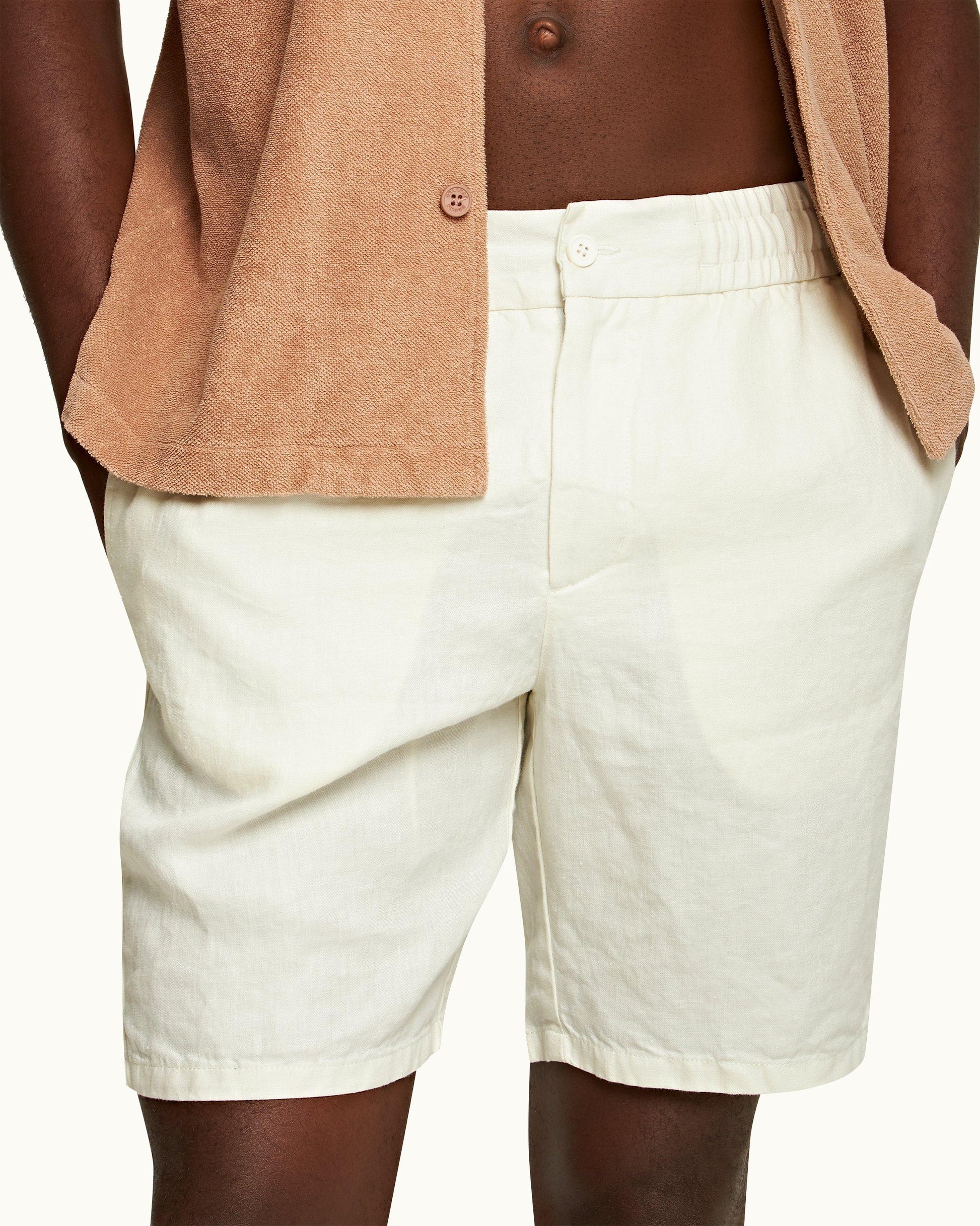 A person wears Orlebar Brown Cornell Linen Shorts in white with an elastic waistband, paired with a beige shirt, hands in pockets, shown from waist to mid-thigh.